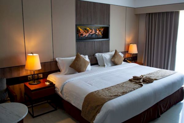 A cozy hotel room with a large bed, decorative pillows, bedside lamps, and a window with curtains. A decorative piece hangs above the bed.