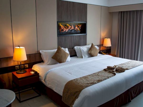A cozy hotel room with a large bed, decorative pillows, bedside lamps, and a window with curtains. A decorative piece hangs above the bed.