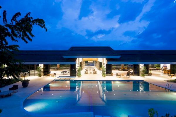 The image features a luxurious, well-lit building with an expansive swimming pool in the foreground against a blue evening sky.