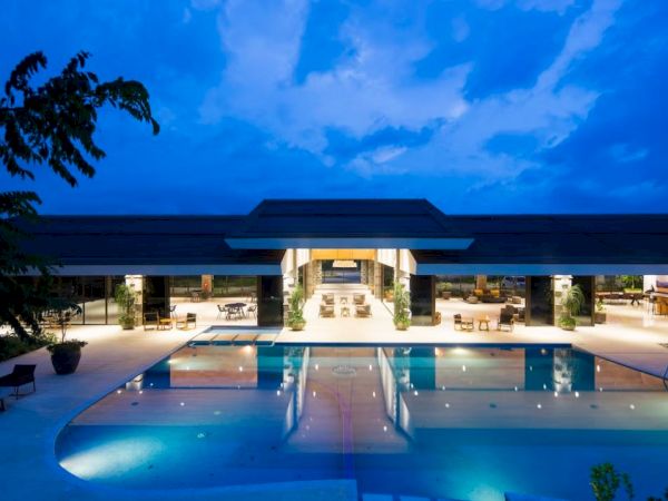 The image features a luxurious, well-lit building with an expansive swimming pool in the foreground against a blue evening sky.
