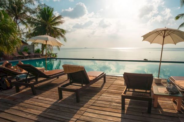 Poolside view with lounge chairs, umbrellas, and lush trees, overlooking a calm sea under a bright sky with scattered clouds.