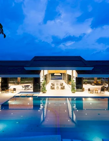 A luxurious modern building with an illuminated pool and outdoor seating, set against a dramatic blue evening sky.