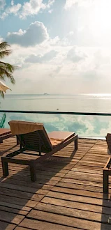 A serene poolside setting with lounge chairs, umbrellas, and a view of the ocean under a partly cloudy sky.