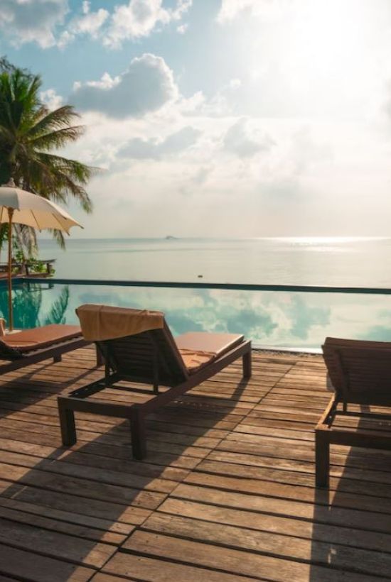A serene poolside scene with lounge chairs and umbrellas overlooking the ocean, framed by palm trees and a sunny sky.