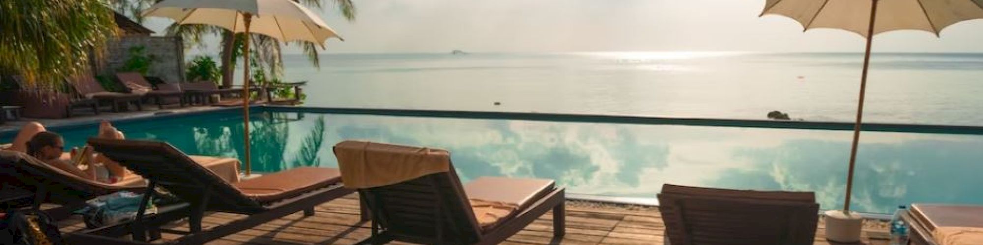 A serene poolside scene with lounge chairs, umbrellas, and palm trees overlooking a calm ocean under a partly cloudy sky.