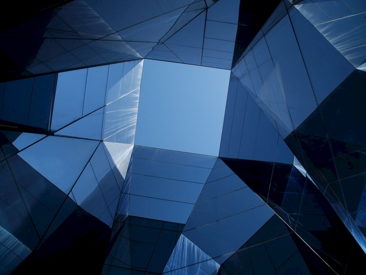The image shows a view of a blue sky through a geometric arrangement of mirrored, angular glass structures.