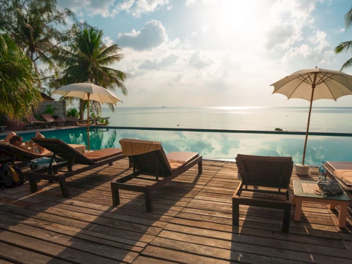 A tranquil poolside scene with lounge chairs, palm trees, umbrellas, and a view of the ocean under a bright sky.
