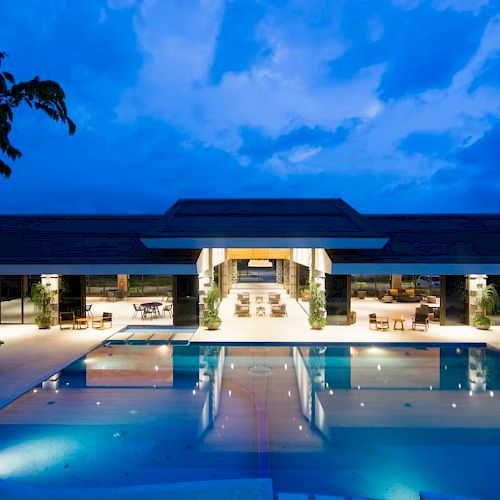 The image shows a modern building with large glass windows and a pool in the foreground, set against a twilight sky.