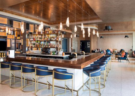The image shows a modern bar with high chairs, pendant lighting, and a well-stocked liquor display, set in a sleek, contemporary space.