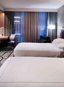 A modern hotel room with two beds, wall-mounted TV, desk, chair, and stylish decor, illuminated by lamps and natural light.