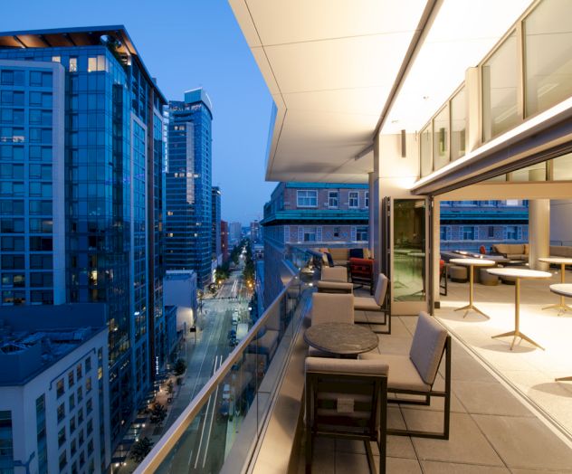 A rooftop terrace with seating overlooks a city street lined with modern buildings, captured at dusk.