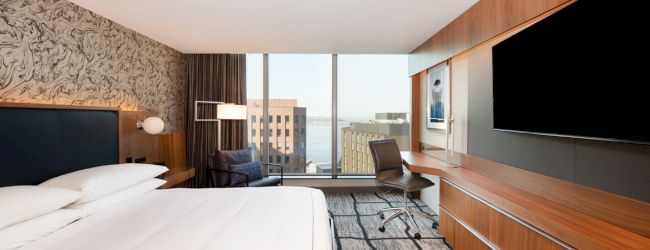 A modern hotel room features a large bed, desk, chair, wall-mounted TV, and window with a city and water view.