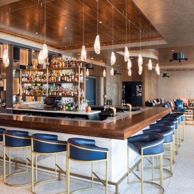 The image shows a modern bar with blue barstools, hanging lights, and a well-stocked back counter, featuring a spacious lounge area.