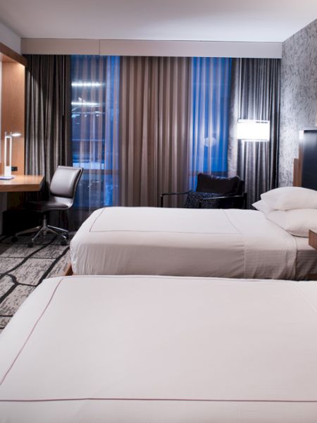 The image shows a modern hotel room with two beds, a wall-mounted TV, a desk with a chair, and stylish decor, illuminated by soft lighting.