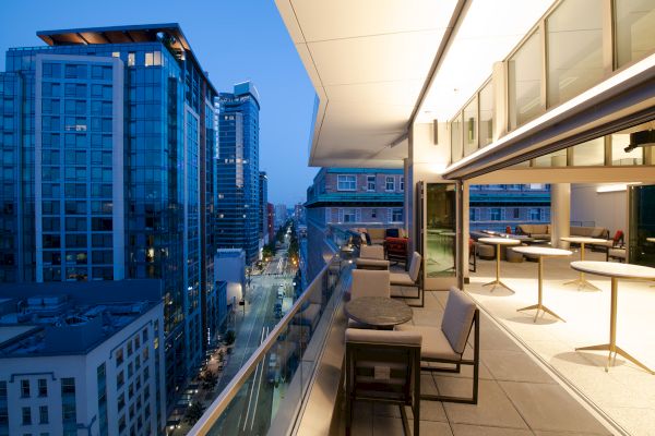 A modern cityscape at twilight with a sleek balcony overlooking tall buildings, featuring outdoor seating and bright lighting inside.