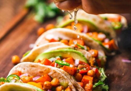 The image shows a hand squeezing lime over tacos filled with chickpeas, avocado, and vegetables, arranged on a wooden board.