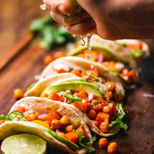 The image shows a hand squeezing lime over tacos filled with chickpeas, avocado, and vegetables, arranged on a wooden board.