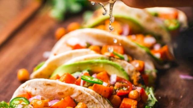 The image shows a hand squeezing lime over tacos filled with chickpeas, avocado, and vegetables, arranged on a wooden board.