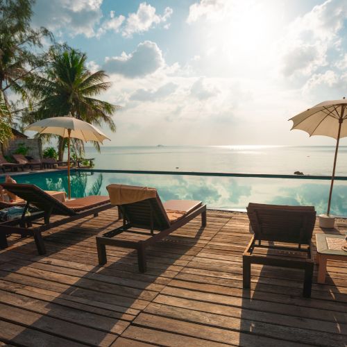 A tropical poolside scene with lounge chairs, umbrellas, and palm trees overlooking a calm sea under a partly cloudy sky.
