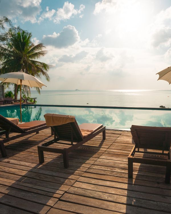 A tropical poolside scene with lounge chairs, umbrellas, and palm trees overlooking a calm sea under a partly cloudy sky.