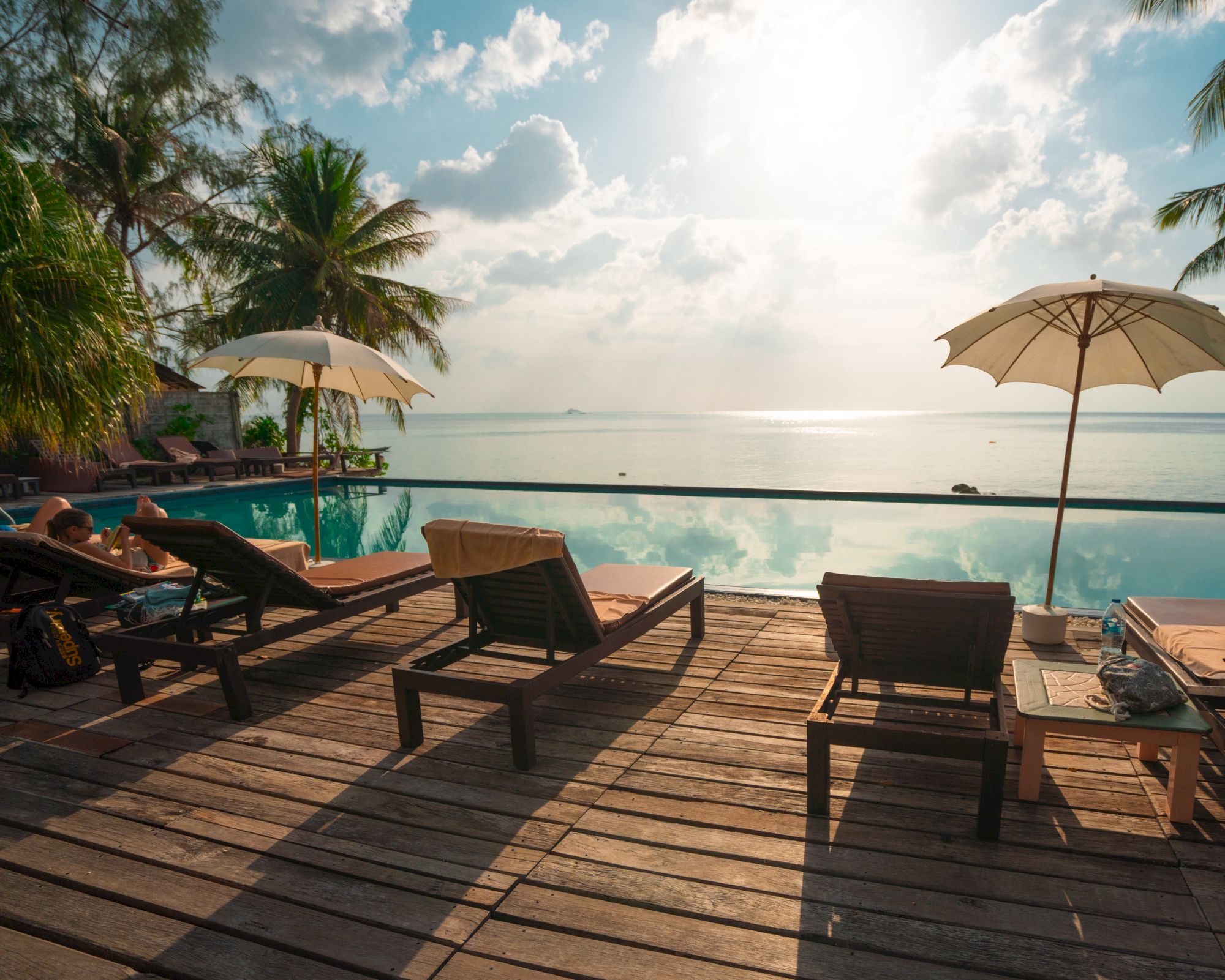 Poolside deck with lounge chairs, umbrellas, and palm trees overlooking a serene ocean view under a cloudy sky.