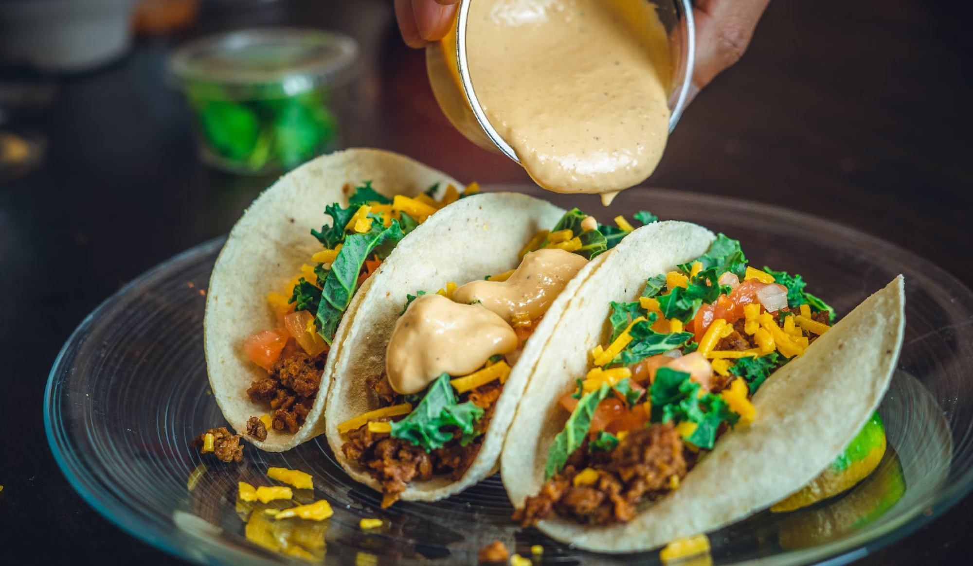 Three tacos are being topped with a creamy sauce, containing ground meat, cheese, lettuce, and tomatoes on a plate.