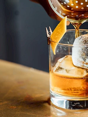 A cocktail is being poured into a glass with ice and a lemon twist garnish, set on a wooden surface.