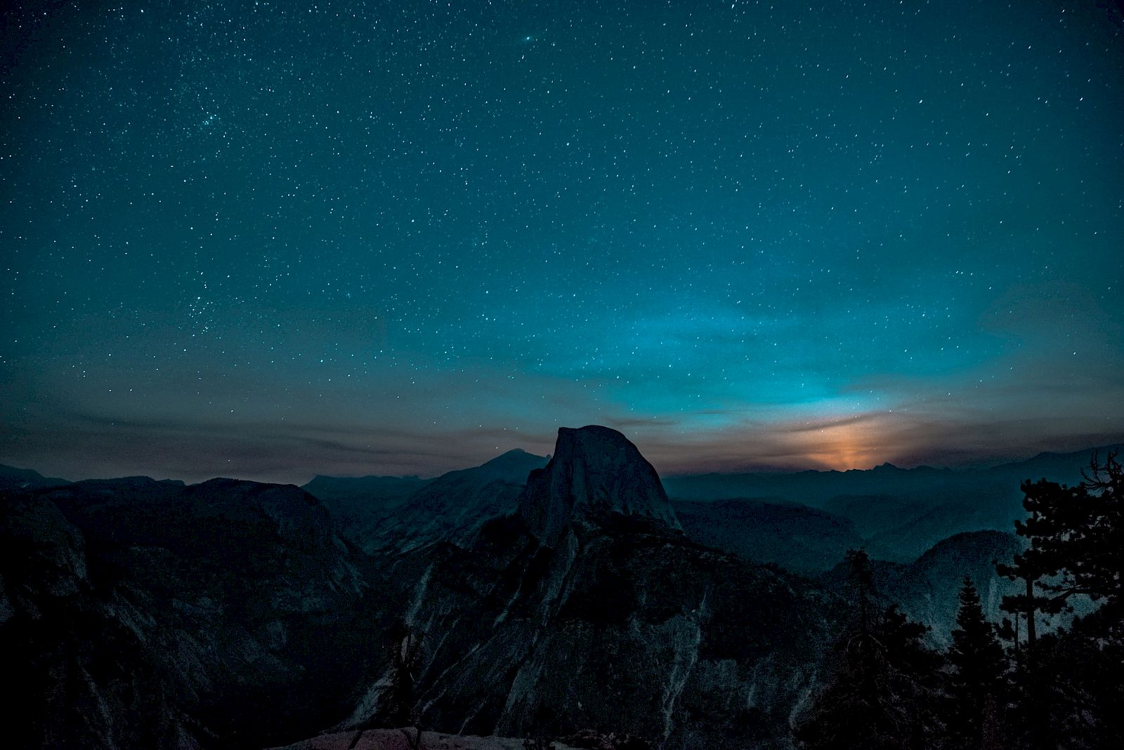 A starry night sky above a rugged mountain landscape with a glowing horizon and silhouetted trees in the foreground.