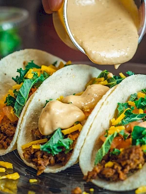 Three tacos filled with meat, cheese, lettuce, and sauce being poured on top, served on a plate.