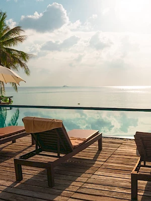 The image shows a serene poolside with loungers, umbrellas, and a view of the ocean under a cloudy sky, surrounded by palm trees.