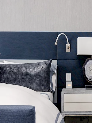 The image shows a modern bedroom with a navy headboard, bedside table, lamp, and decorative pillows, creating a stylish aesthetic.