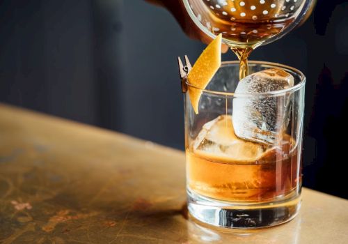 A glass with an amber liquid and a large ice cube is being poured into, garnished with a twist of citrus peel on the rim.