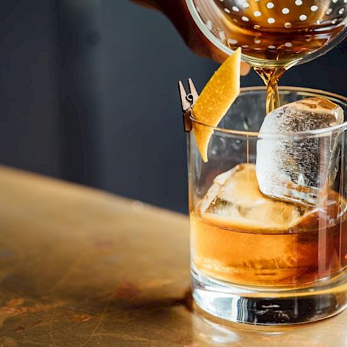 A glass with an amber liquid and a large ice cube is being poured into, garnished with a twist of citrus peel on the rim.