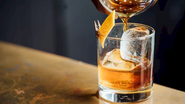 A glass with an amber liquid and a large ice cube is being poured into, garnished with a twist of citrus peel on the rim.