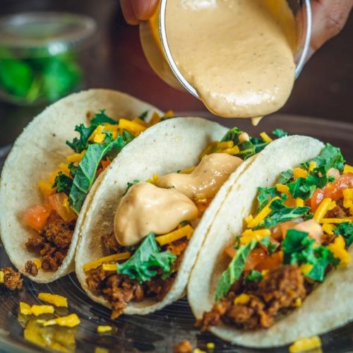 Three tacos filled with beef, lettuce, tomatoes, and cheese on a plate, topped with creamy sauce being poured over them.
