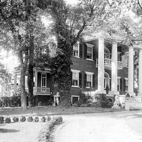 The image shows a large, historic building with tall columns and a wrap-around porch surrounded by trees and landscaped gardens.
