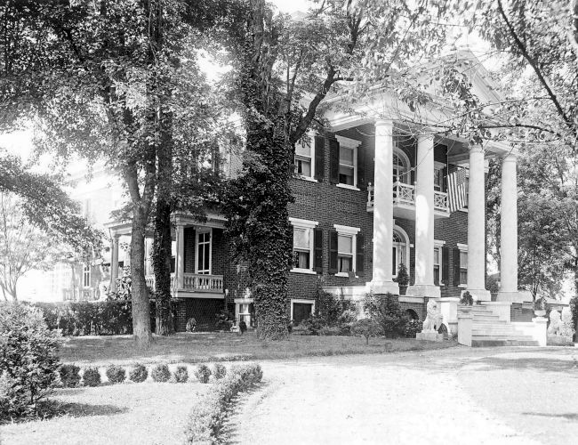 The image shows a large, historic building with tall columns and a wrap-around porch surrounded by trees and landscaped gardens.