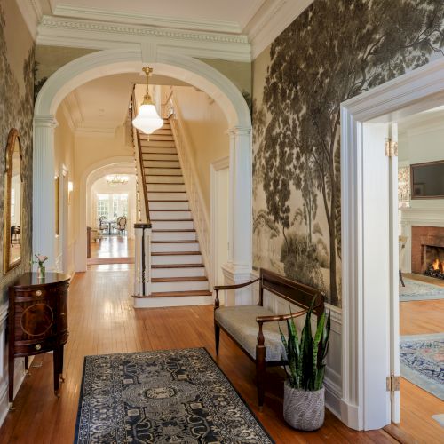 A bright hallway with framed arches, a staircase ahead, a rug, potted plant, hallway table, and a doorway to a living room with a fireplace.