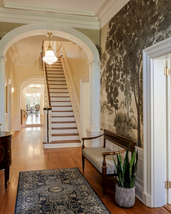A bright hallway with framed arches, a staircase ahead, a rug, potted plant, hallway table, and a doorway to a living room with a fireplace.