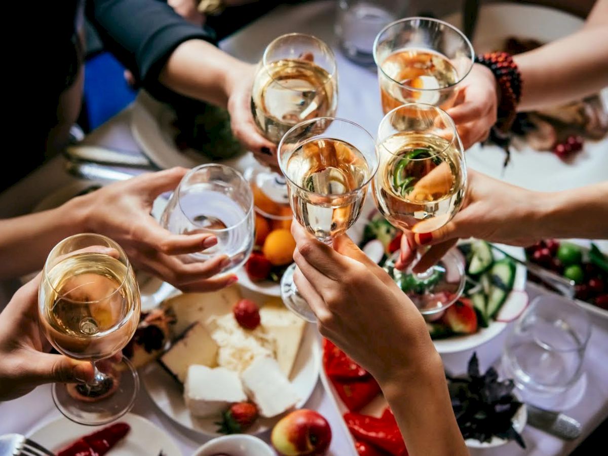 A group of people clinking glasses filled with white wine and water over a table with various appetizers and fruits.