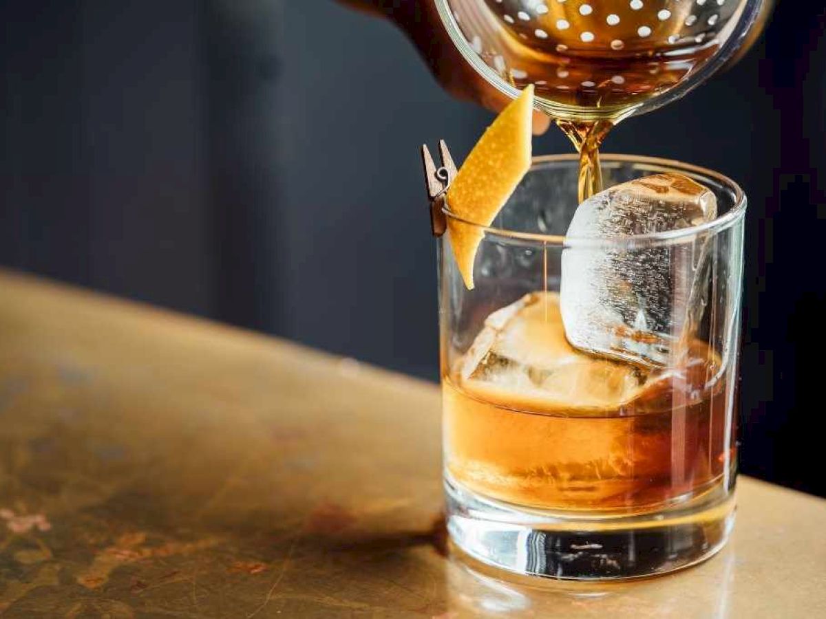 A person is pouring a brown beverage into a glass with ice and an orange peel garnish on a wooden surface.