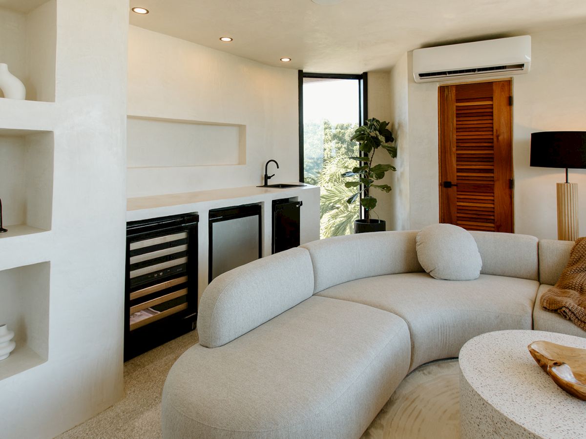 A modern living room with a curved sofa, built-in shelves, kitchenette, and minimal decor, featuring neutral tones and soft lighting.