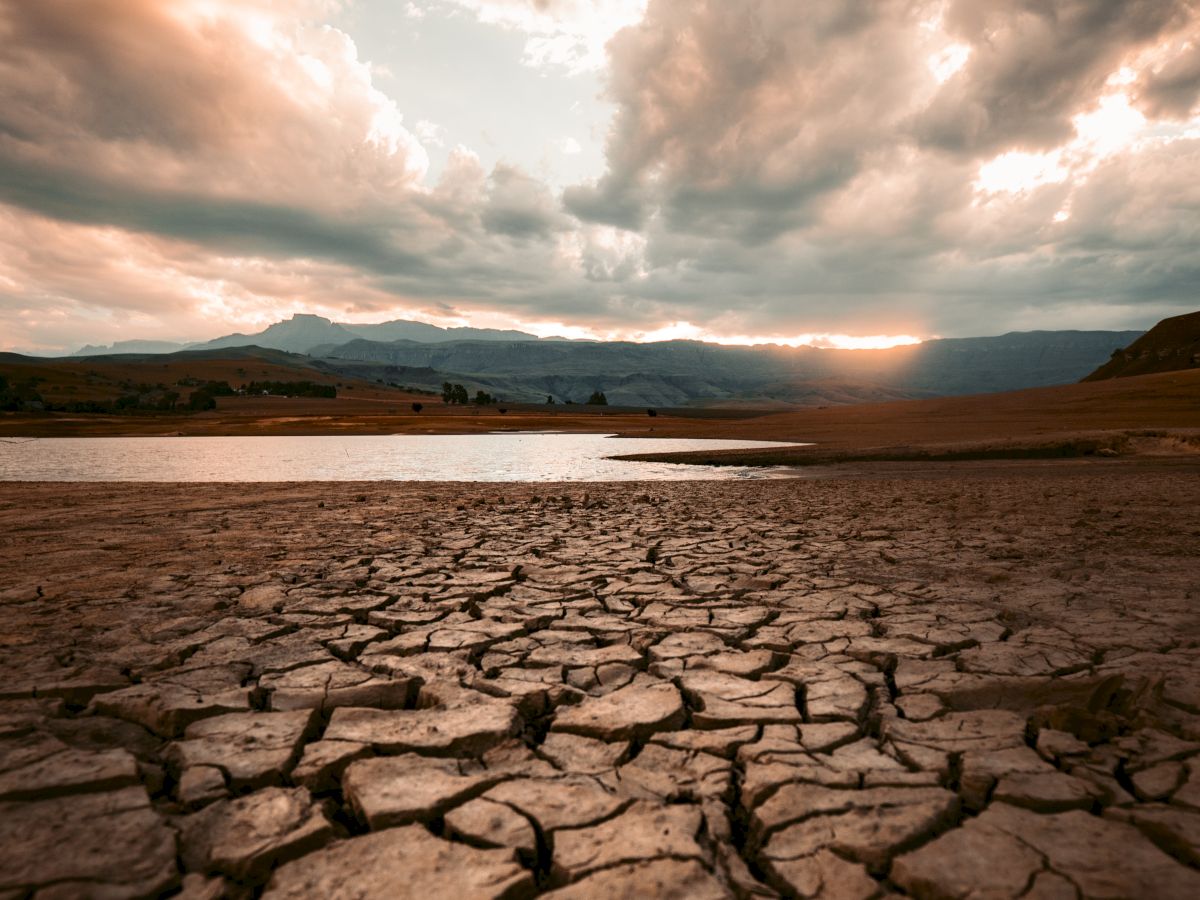 A cracked, dry landscape under a dramatic cloudy sky with a distant view of water and mountains in the background.