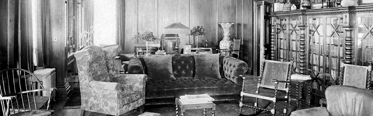 A vintage living room with ornate wooden walls, upholstered chairs, a couch, a cabinet with glass doors, and various decorations displayed.