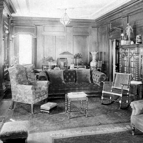 A vintage living room with ornate wooden walls, upholstered chairs, a couch, a cabinet with glass doors, and various decorations displayed.