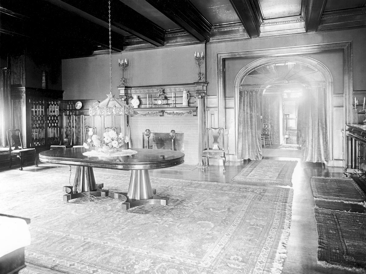The image shows a vintage room with a large area rug, a round table, and a fireplace, featuring ornate woodwork and a chandelier.