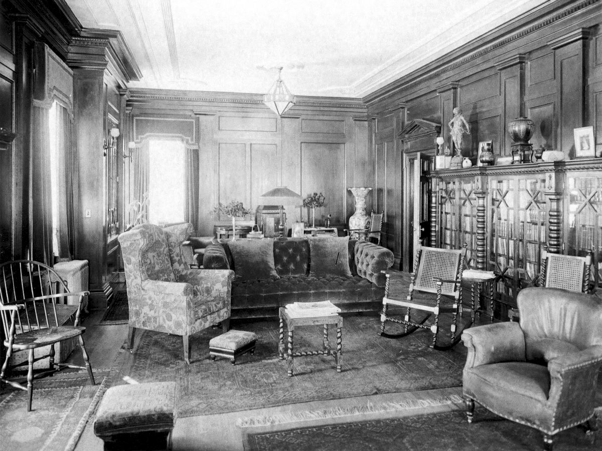 A vintage living room with ornate wood paneling, a variety of chairs, a bookshelf, and decorative items on the mantle and shelves.