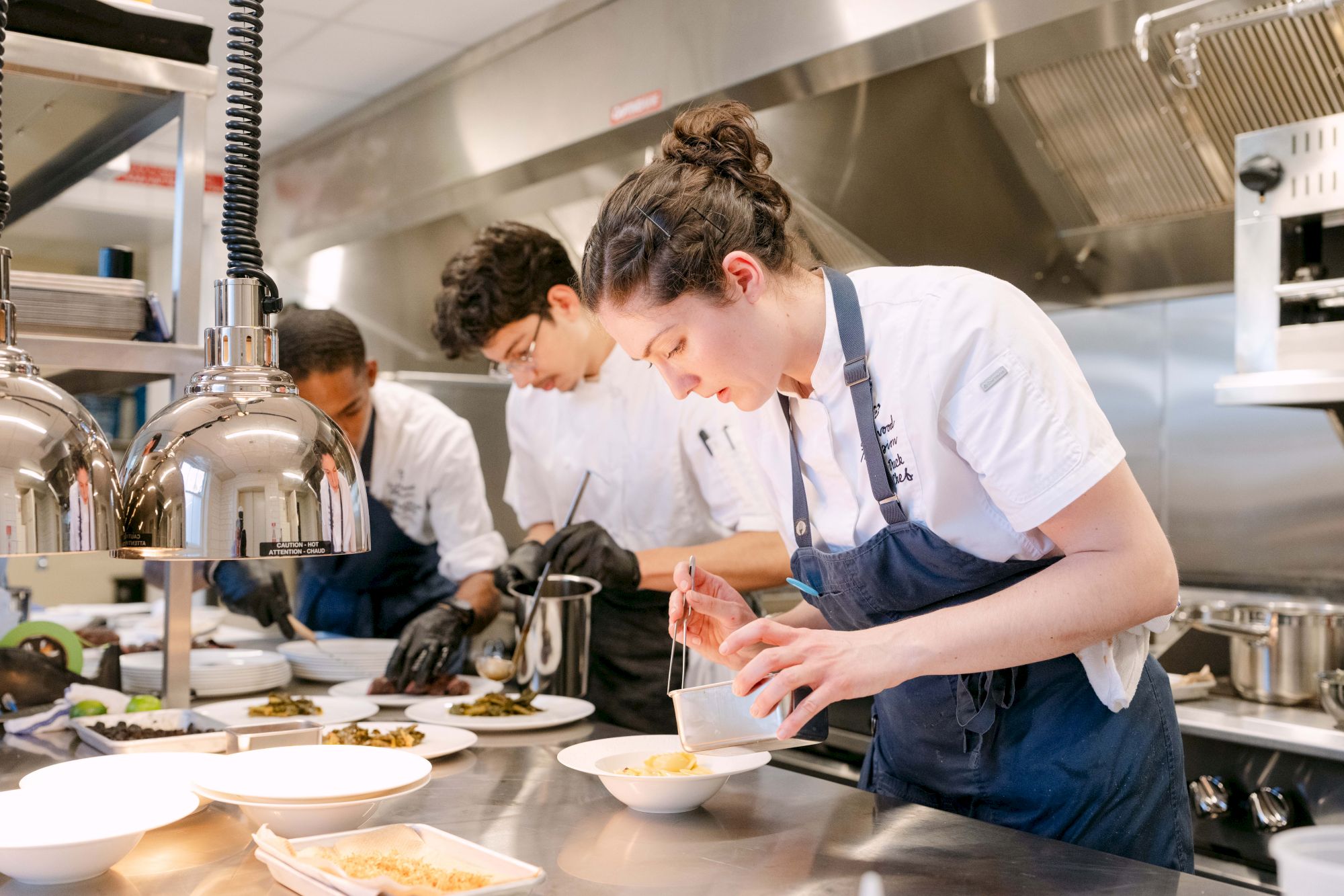 忙しい kitchen with students in white shirts and aprons plating dishes and tasting sauces, focused on precision and teamwork.