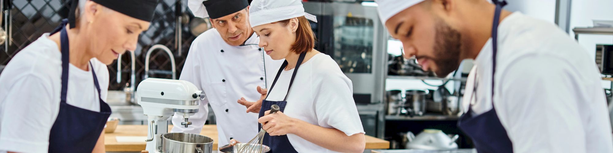 Chefs in a kitchen are working with baking equipment, wearing white shirts, black aprons, and caps, focused on preparing food.