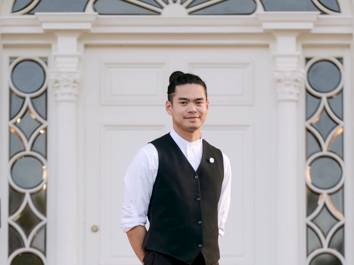 A stylish man in a white shirt and black vest stands casually in front of a white, elegant door with circular window panels, posing confidently.